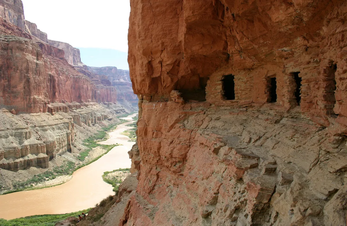 Small stone rooms with dark doorways built into a red cliff face, high above a muddy river.