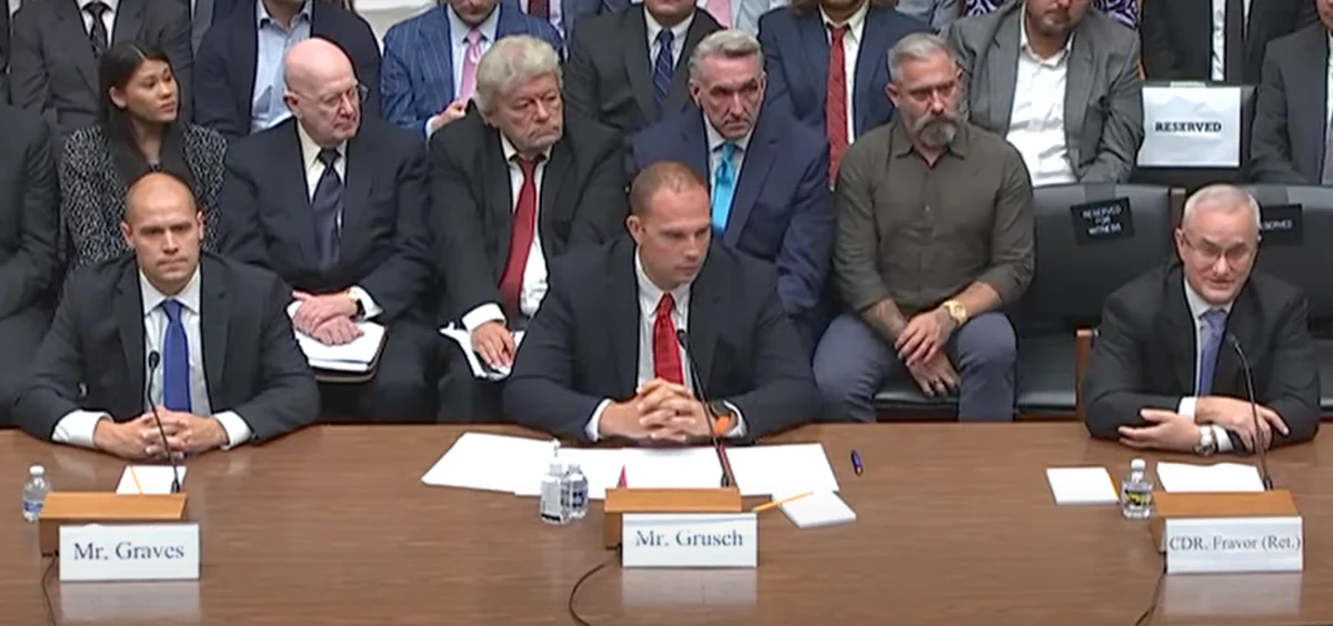 Three men in dark suits at a witness table with name plates for Mr. Graves, Mr. Grusch and Cdr. Fravor, with spectators seated behind them.
