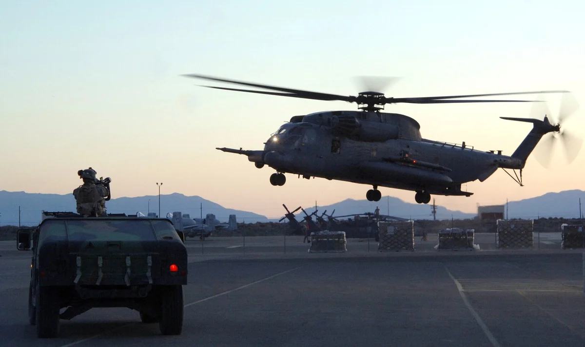 A large military helicopter lands at dusk on an airfield ramp while an airman in uniform stands on an armoured vehicle in the foreground, mountains behind.