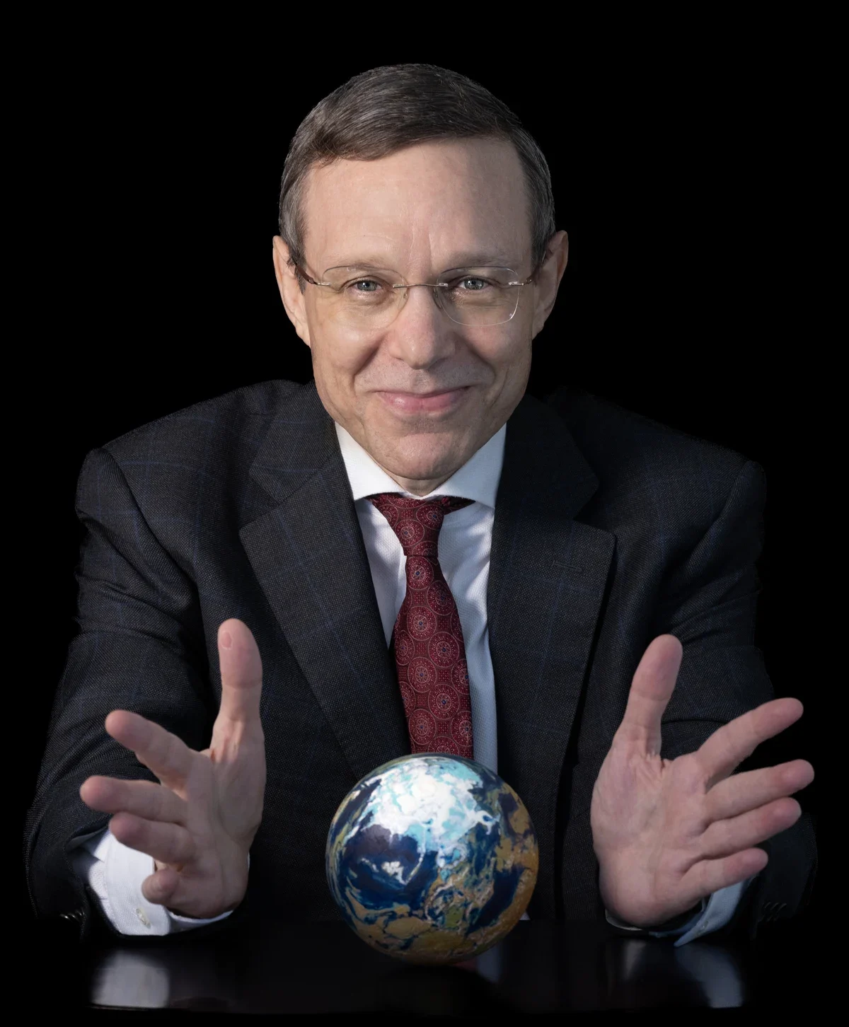 A man with short grey hair in a dark jacket, smiling, photographed against a plain background.