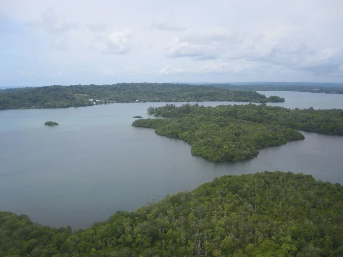 A green tropical coastline on Manus Island under a cloudy sky, with calm water in the foreground.