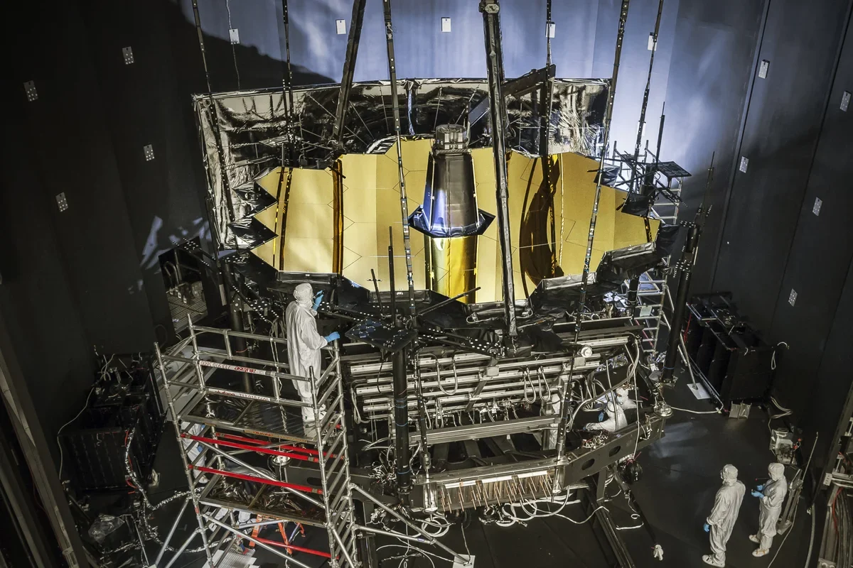 Engineers in white clean-room suits stand on a lift beside the large gold honeycomb mirror of the Webb telescope inside a test chamber.