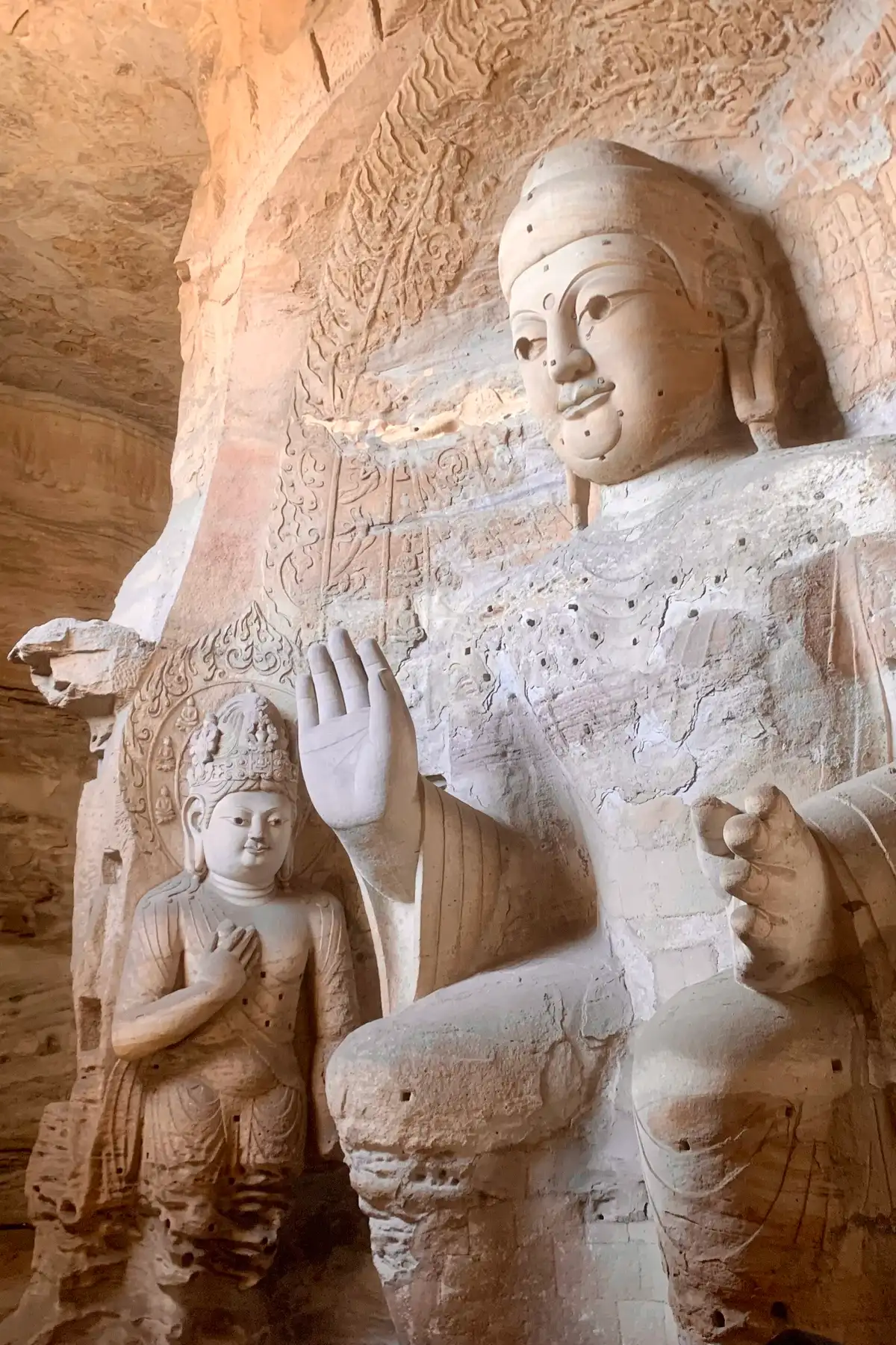 A colossal seated stone Buddha with one hand raised, a smaller attendant figure at its side, in a sandstone cave.