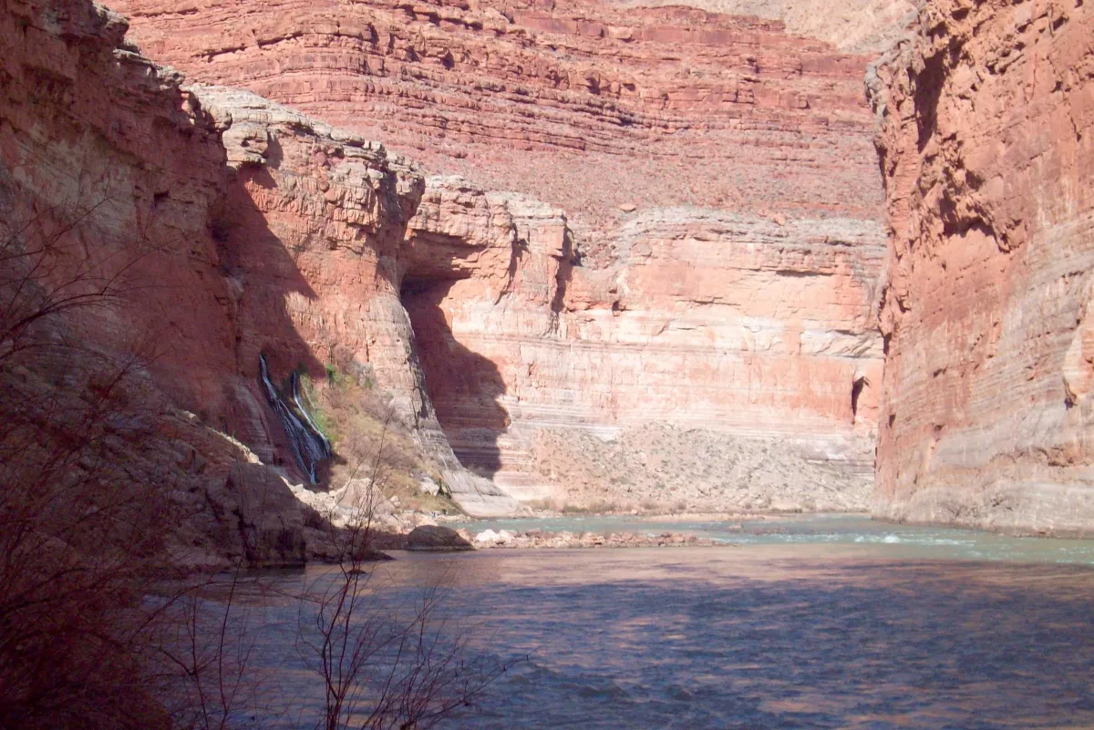 A river below tall banded red cliffs, with green growth at the foot.