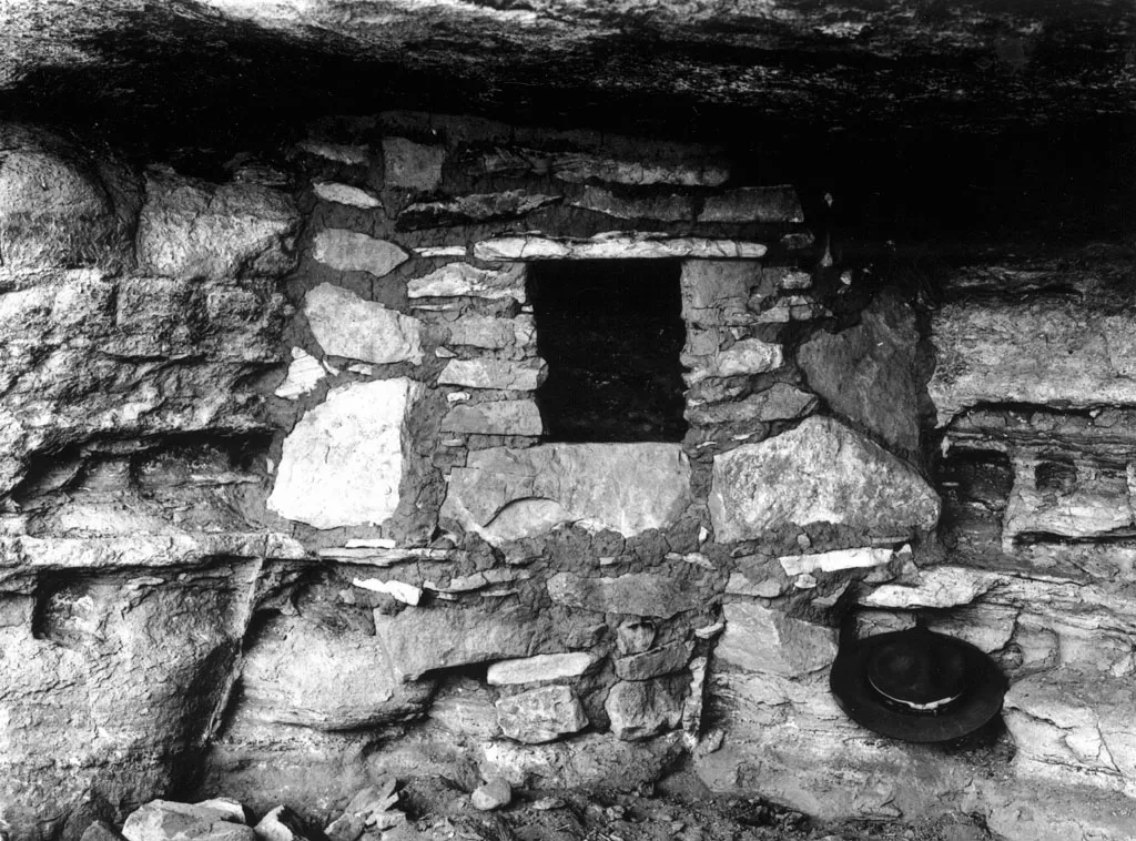 A black and white photograph of a small stone wall with a dark square doorway, built beneath an overhanging ledge.
