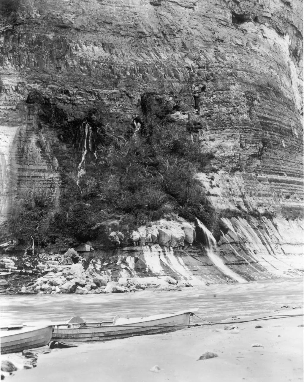 A black and white photograph of boats on a beach below a high cliff streaked with springs.
