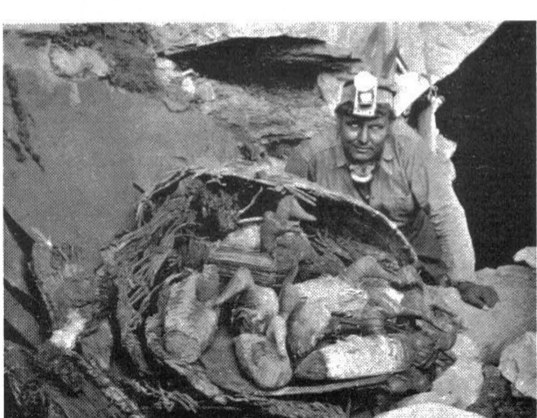 Black and white photograph inside the cave: a man in a miner's headlamp behind an opened basket holding a pile of rush duck decoys and wrappings.
