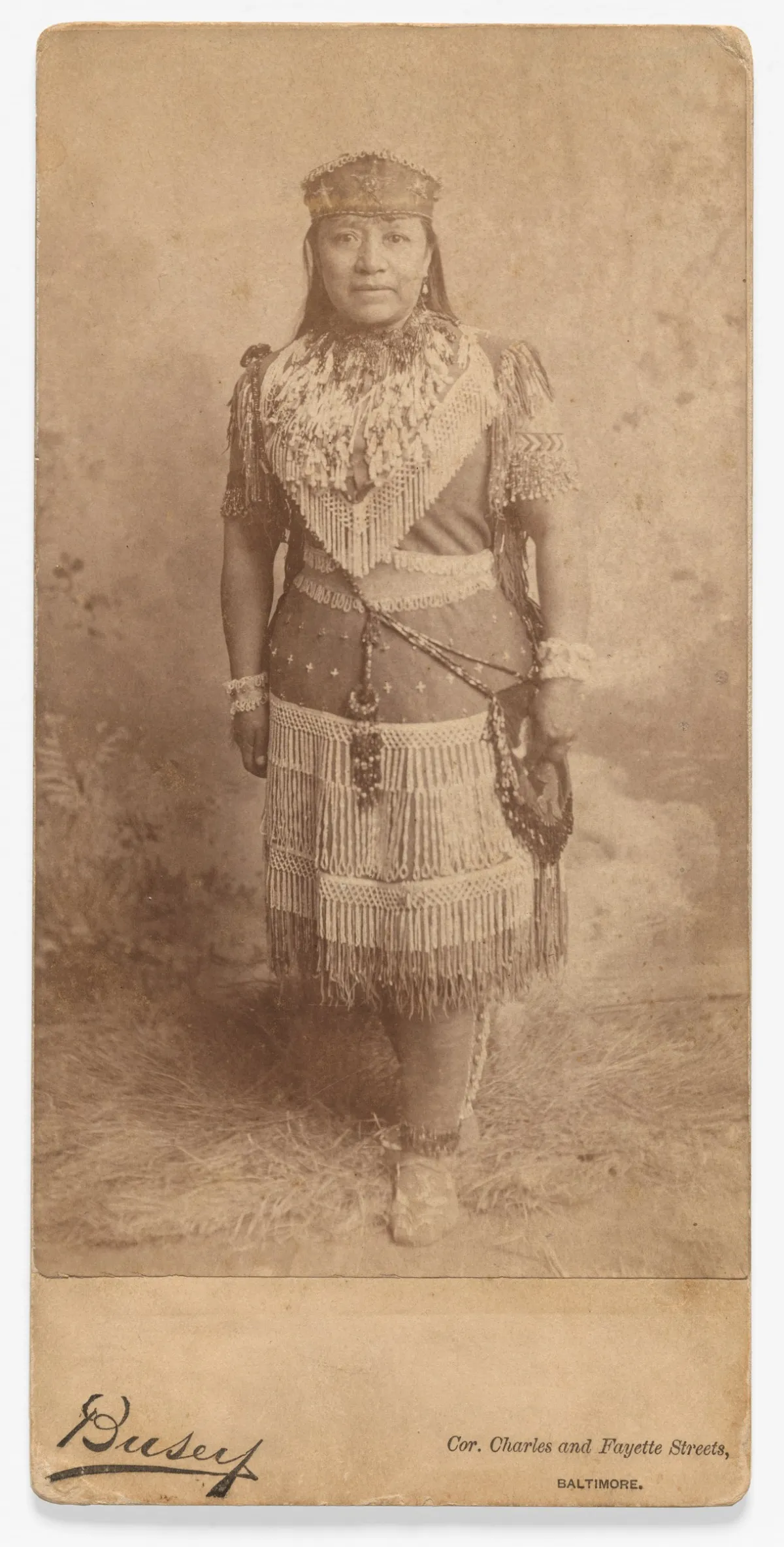 An old studio photograph of a woman standing full length in a fringed and beaded dress with a headdress.