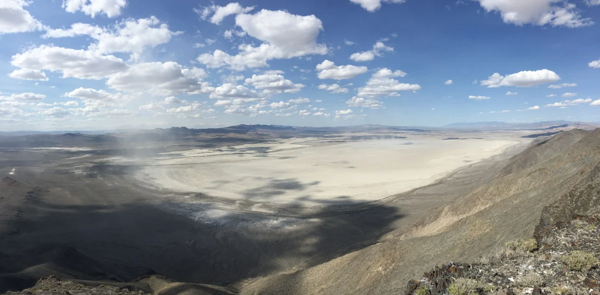 A wide desert basin seen from a mountain, a pale dry lake bed under scattered clouds.