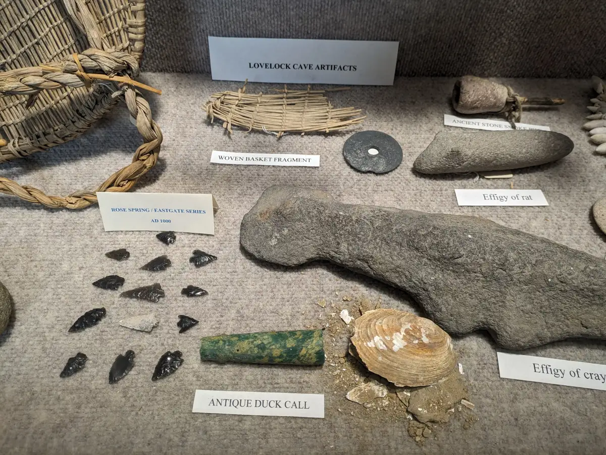 A museum case of objects from Lovelock Cave: a woven basket fragment, stone effigies, obsidian points and a green duck call, with typed labels.