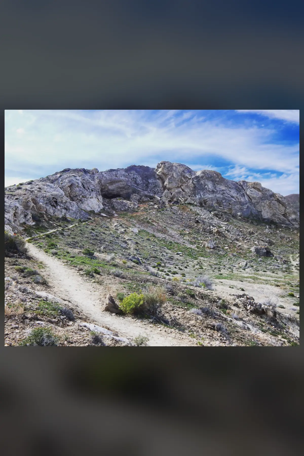 A rugged grey limestone hill under a blue sky with a trail climbing toward a dark cave mouth near the top.