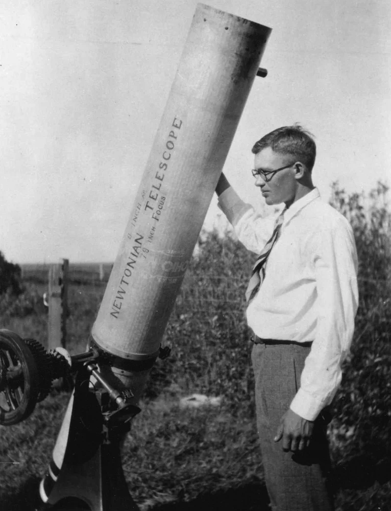 A young man in a white shirt and tie stands beside a tall tube telescope labelled Newtonian Telescope, outdoors by a field