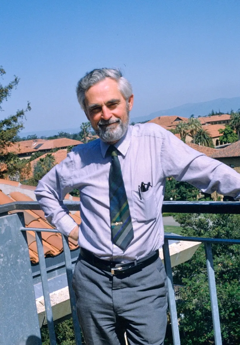 A grey-bearded man in a pale shirt and striped tie leans on a balcony rail, smiling, with red-tiled roofs and hills behind him.