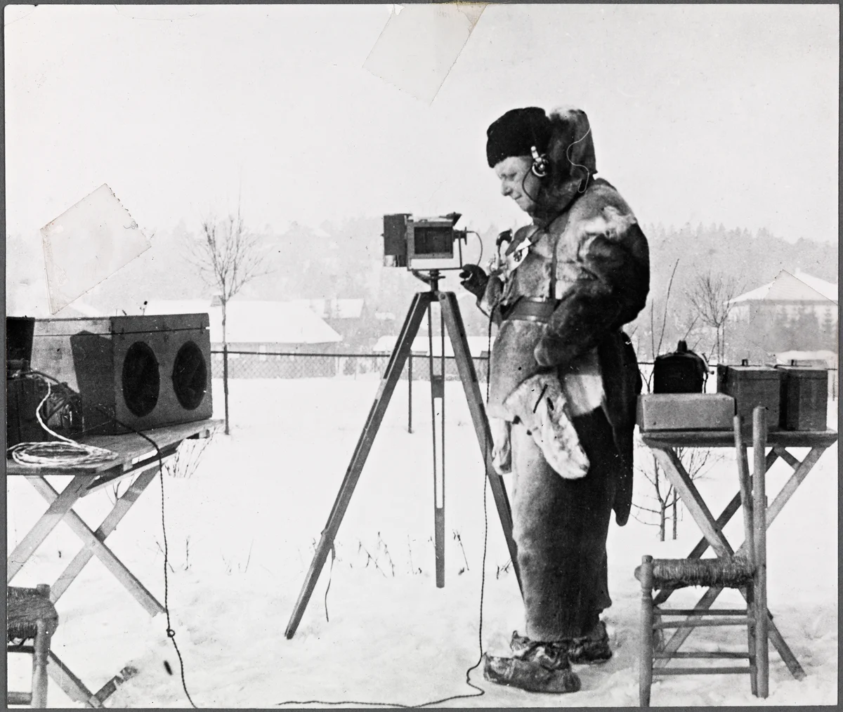A man in a fur coat and hat stands in snow beside a camera on a tripod, with equipment boxes on folding stands around him.