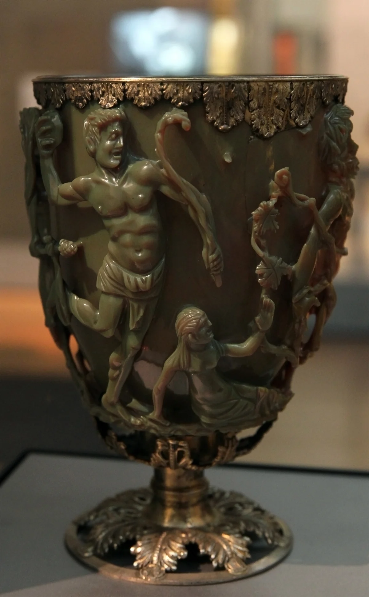 The same glass cup in ordinary light, an opaque olive green, showing a muscular satyr with a raised arm and vine stems on its side.
