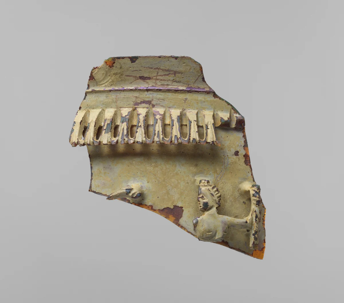 A broken piece of yellowish-green glass with a row of cut teeth along the top and a small carved figure holding a staff at the lower right.