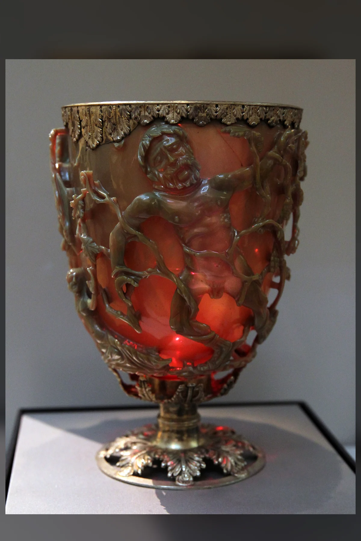 A Roman glass cup on a silver-gilt foot, glowing deep red from a light inside, with a bearded man caught in coiling vines carved in relief on its outer wall.