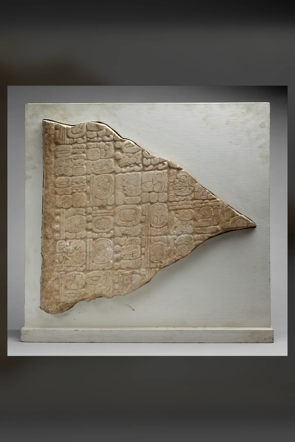 A wedge-shaped broken slab of pale stone carved with rows of square Maya glyph blocks, mounted on a white panel