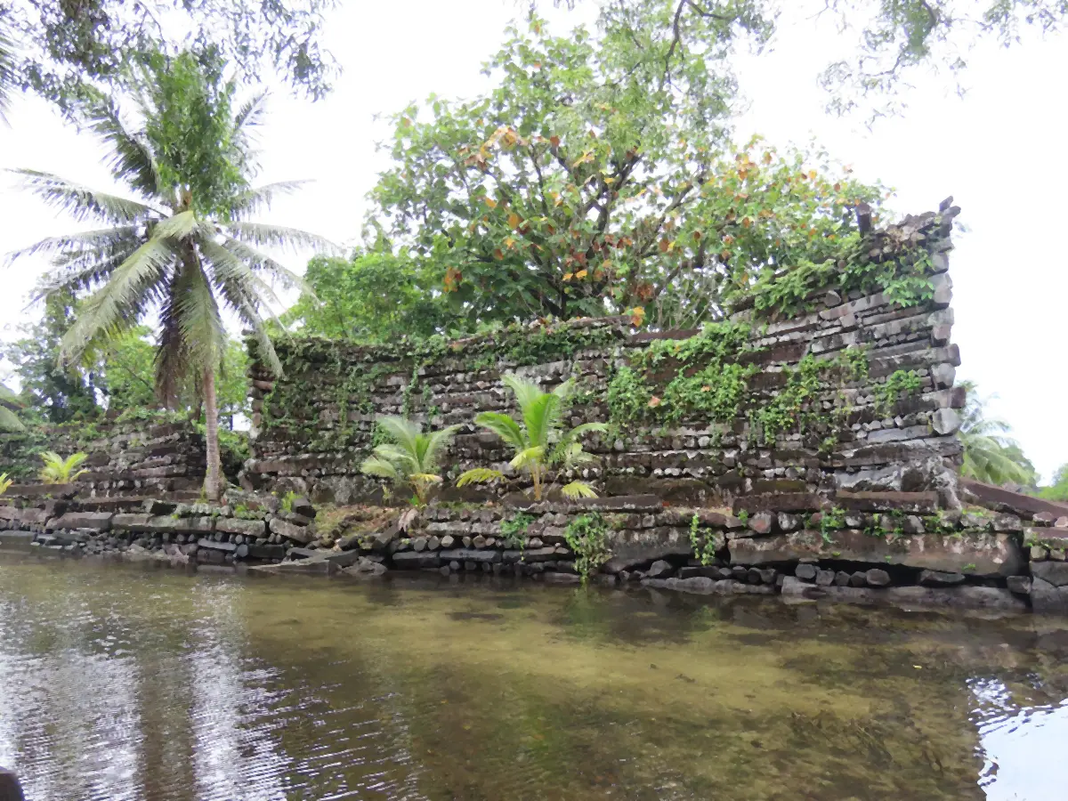 A long basalt wall with an upswept corner above a low stone edge and shallow water, young palms against it.