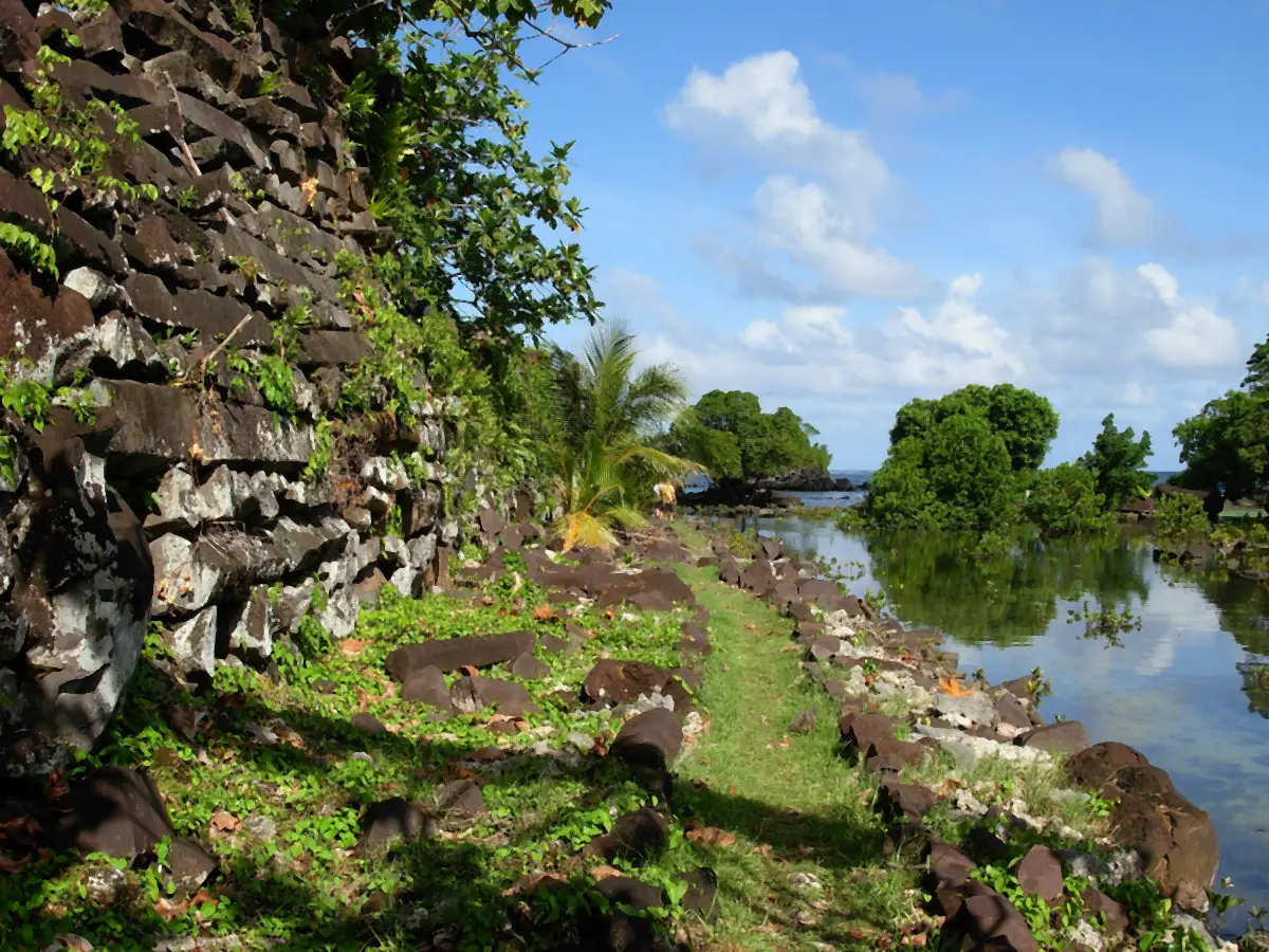 A wall of stacked basalt at left, a grass path, and water with mangrove islets at right.