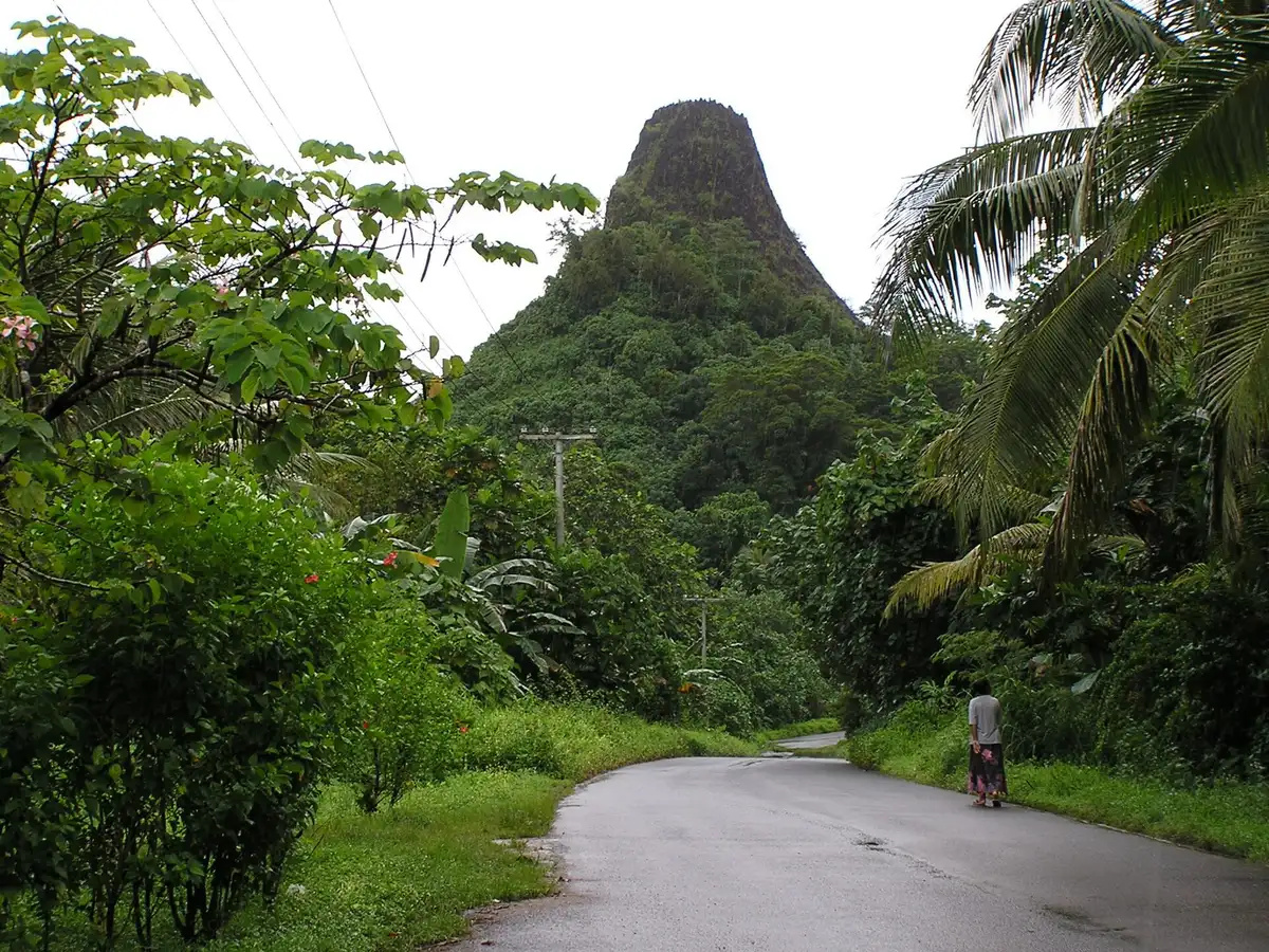 A steep dark volcanic plug rising from green forest above a wet road, with one person walking.