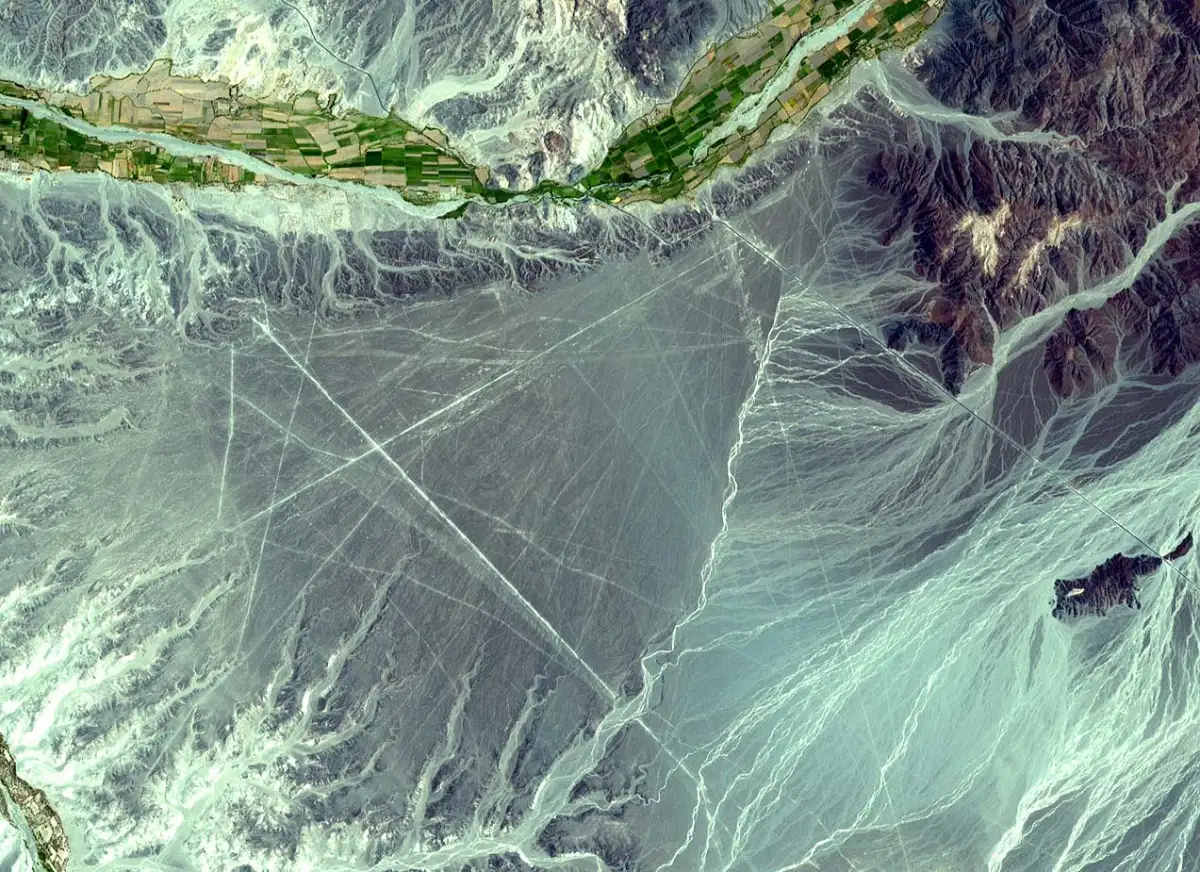 A false-colour satellite view of a desert plain crossed by long straight lines and wedge shapes, a green river valley at the top.