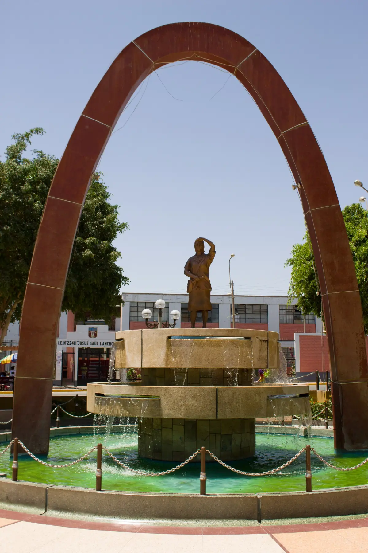 A statue of a woman with raised arm under a tall rust-coloured arch, above a fountain in a town square.