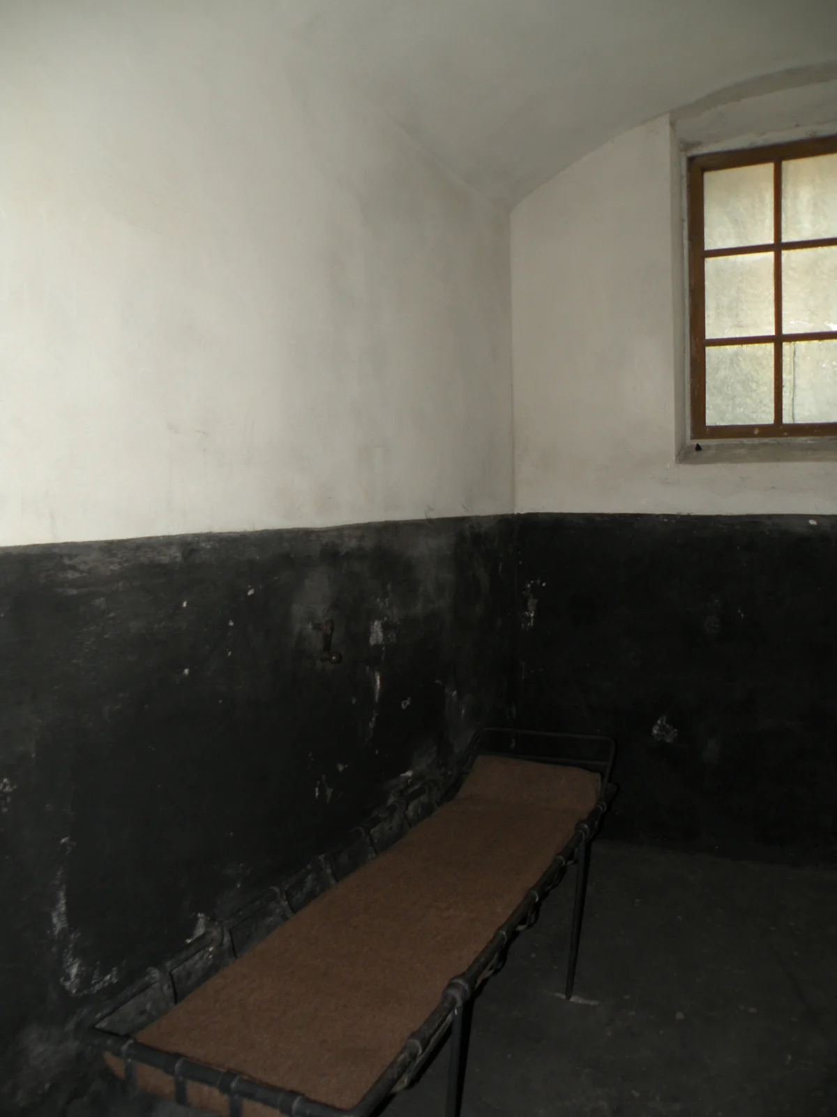 A vaulted prison cell with white upper walls and a black lower band, a window high on one side and a narrow iron bed.