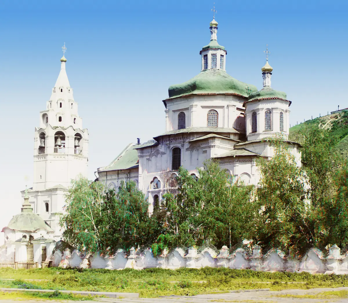 A white church with green domes and a tall white bell tower behind a scalloped wall, under a blue sky.