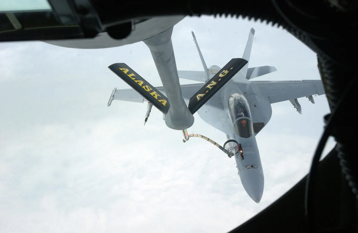 A grey two-seat Super Hornet fighter flies close beneath a tanker's refuelling boom, high over white cloud, seen from the tanker's boom window.