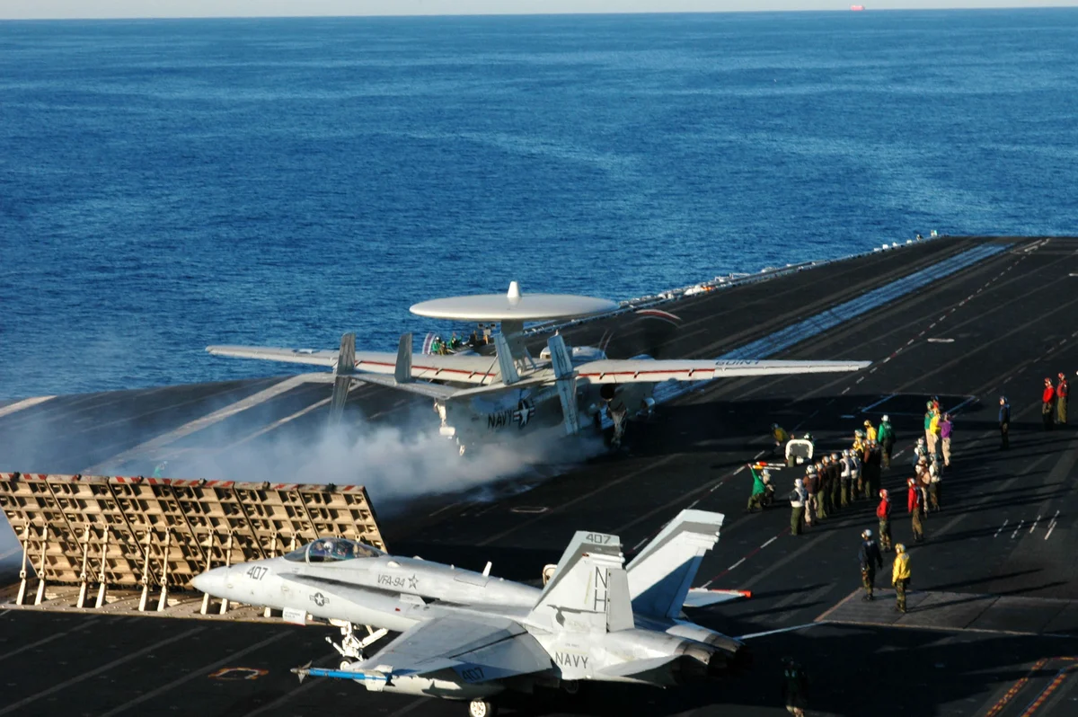 A twin-propeller aircraft with a large radar disc on its back launches from a carrier's catapult in a cloud of steam, as deck crew watch and a fighter waits beside it.
