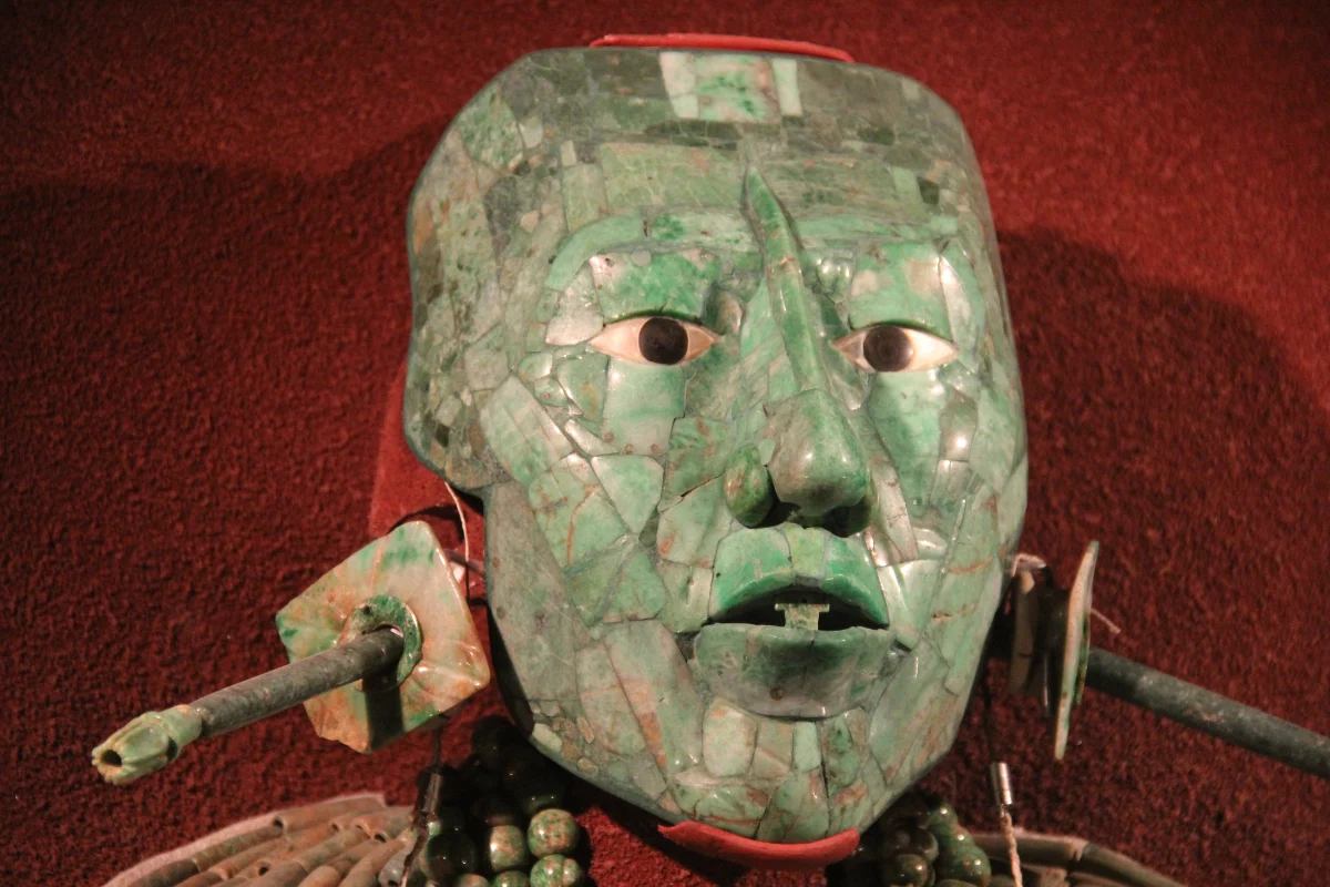 A death mask made of small green jade pieces fitted together, with white shell eyes and dark obsidian pupils, displayed on a red background.
