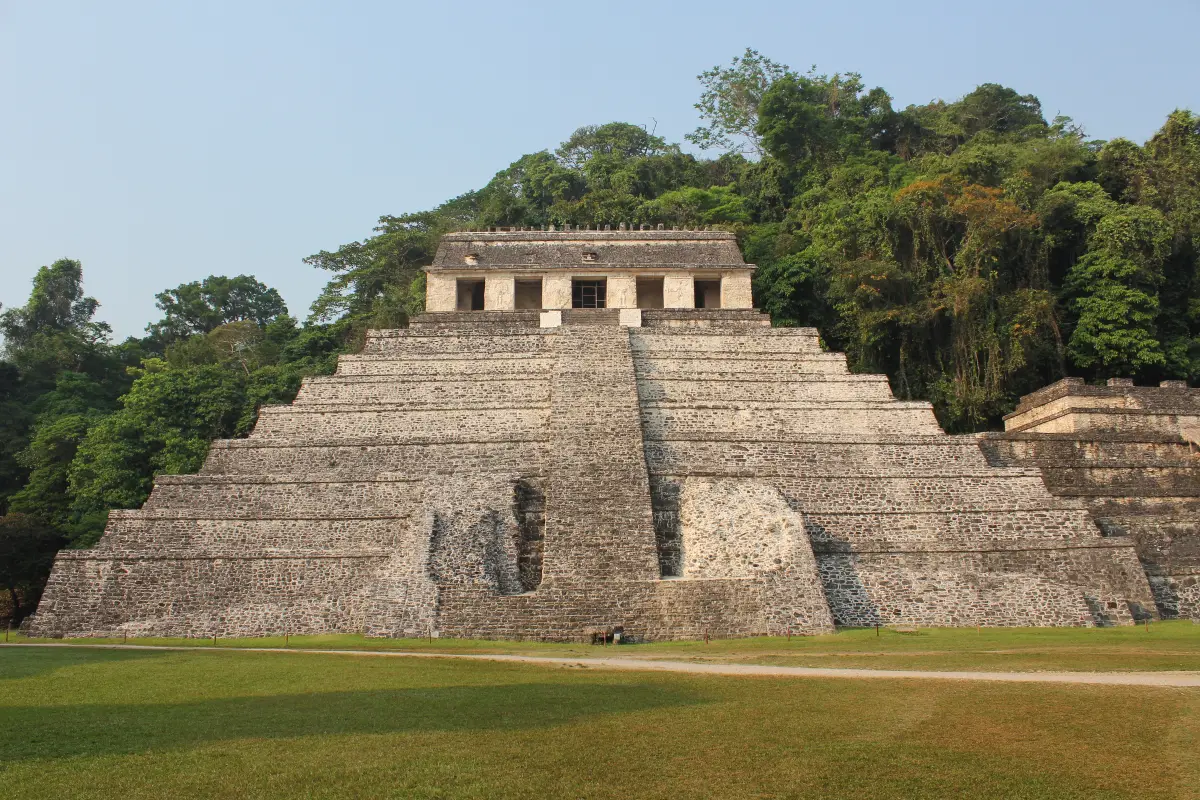A stepped stone pyramid with a long temple on top, rising out of green rainforest on a sunny day.