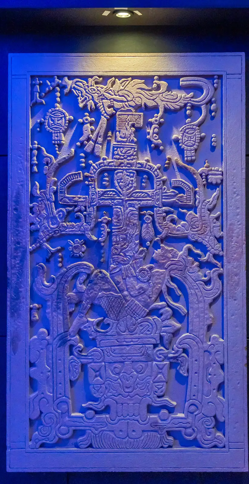 A replica of the lid, its whole carved face seen straight on under blue light: a man reclining with knees bent and hands raised, a tall carved form rising behind him topped by a bird, open jaws below, all inside a border of carved symbols.