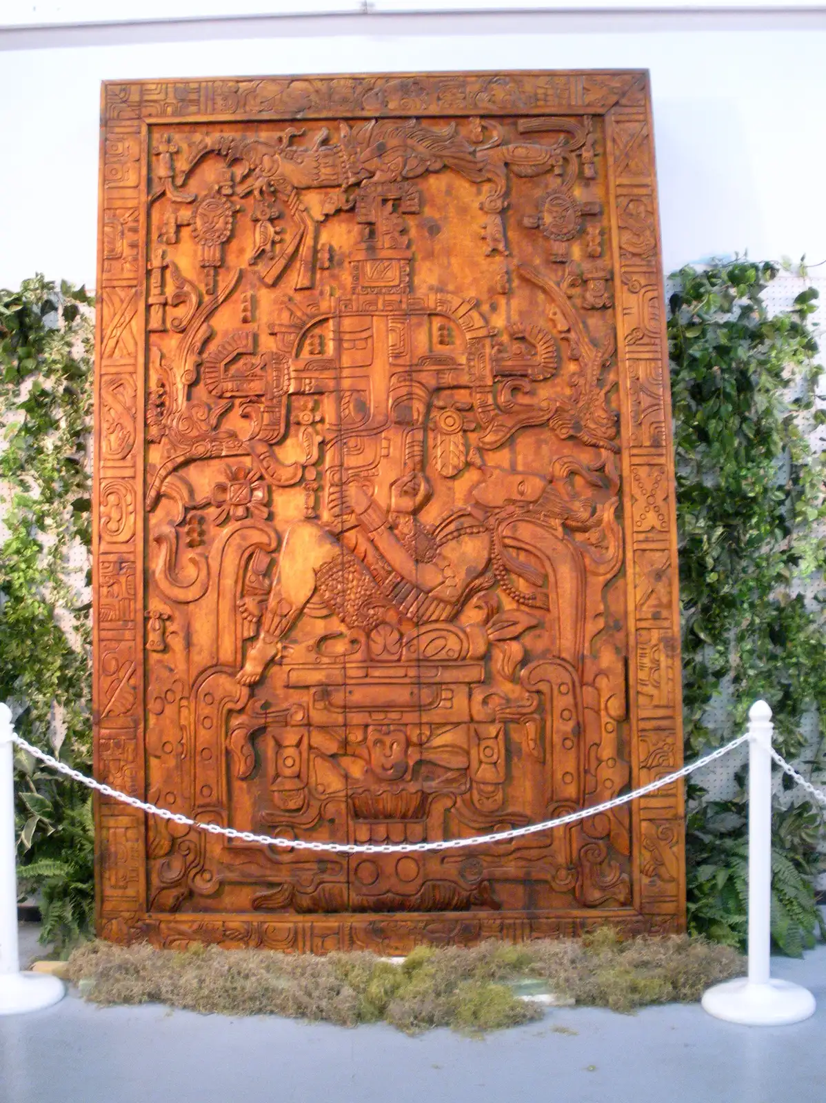 A dark wooden copy of the carved lid displayed upright behind a rope barrier in a museum hall.