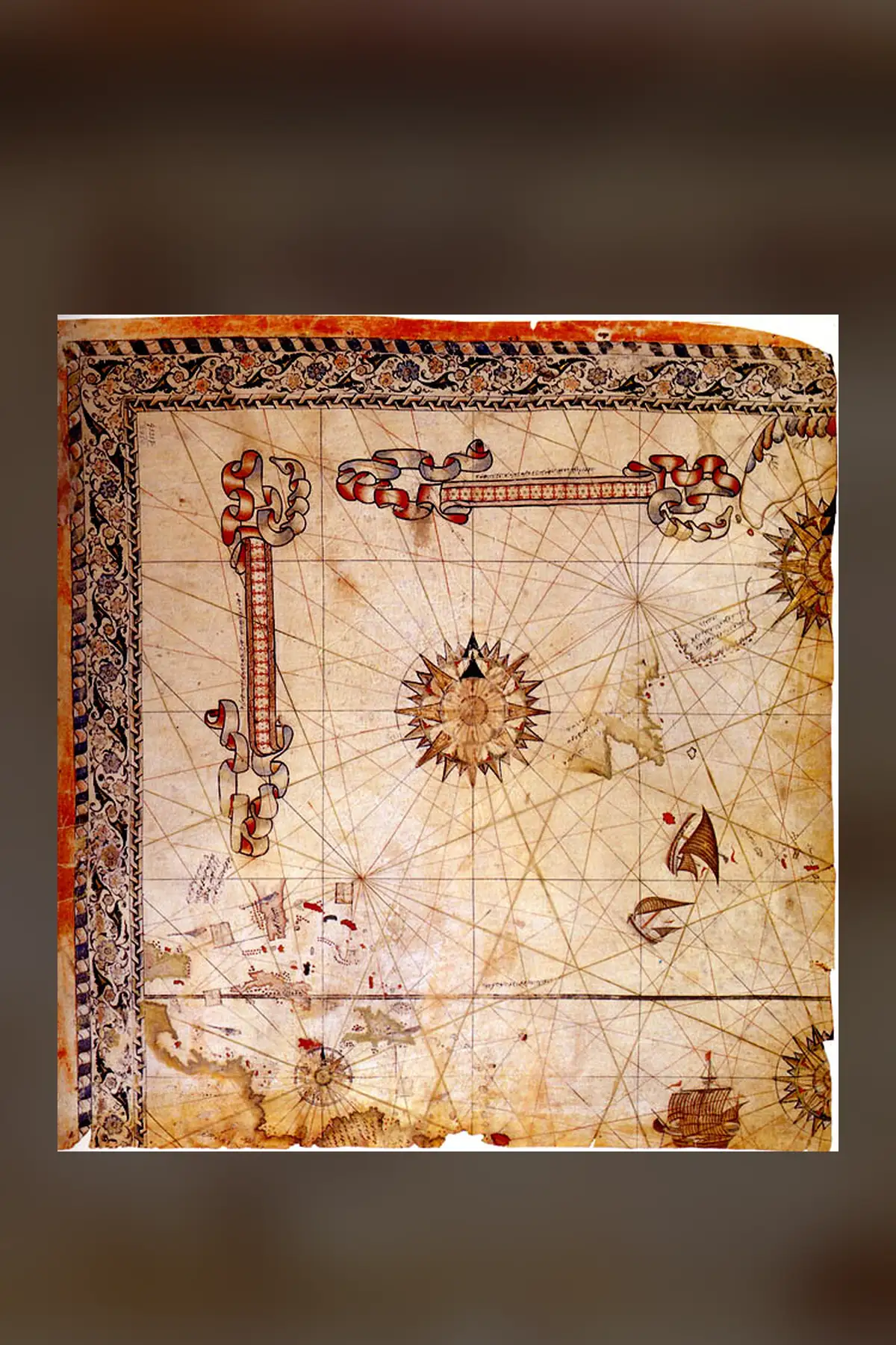 The surviving corner of Piri Reis's world map of 1528: a square parchment crossed by rhumb lines, with a flowered border at the top and left, scale bars in ribbons, compass roses, three ships and the Caribbean islands low on the left.