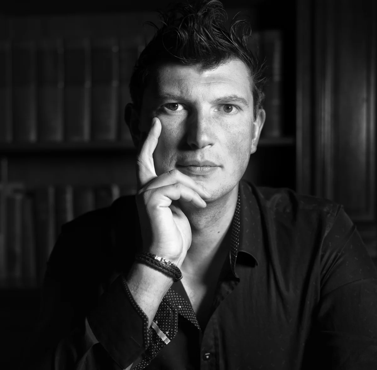 Black and white portrait of Konstantin Batygin seated in front of bookshelves, one hand raised to his face.