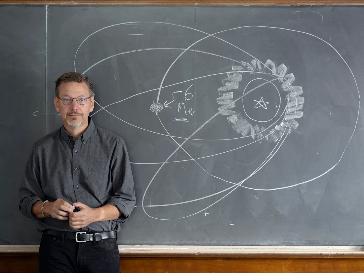 Michael Brown at a chalkboard on which an elongated orbit is drawn, with a planet and the note about six Earth masses beside it.