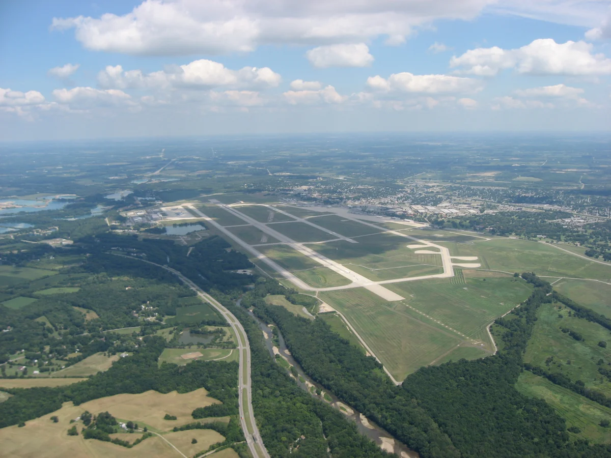 Aerial view of a large airfield with long runways among green fields and woods, a town spreading beyond it to the horizon.