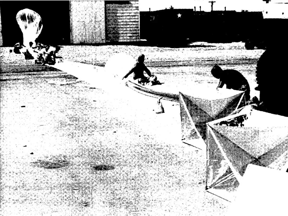 Men on an airfield preparing a balloon launch, with large folded radar reflectors of foil and wood lying on the ground in the foreground.