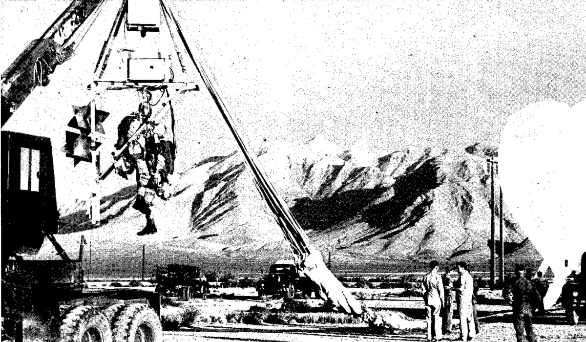 A test dummy in a flight suit hanging from a crane-like launch rig on a truck, with men and a balloon nearby and mountains behind.