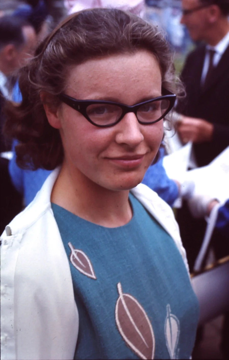 A young woman with dark wavy hair and cat-eye glasses, wearing a teal top with leaf shapes and a white jacket, smiling slightly at an outdoor gathering.
