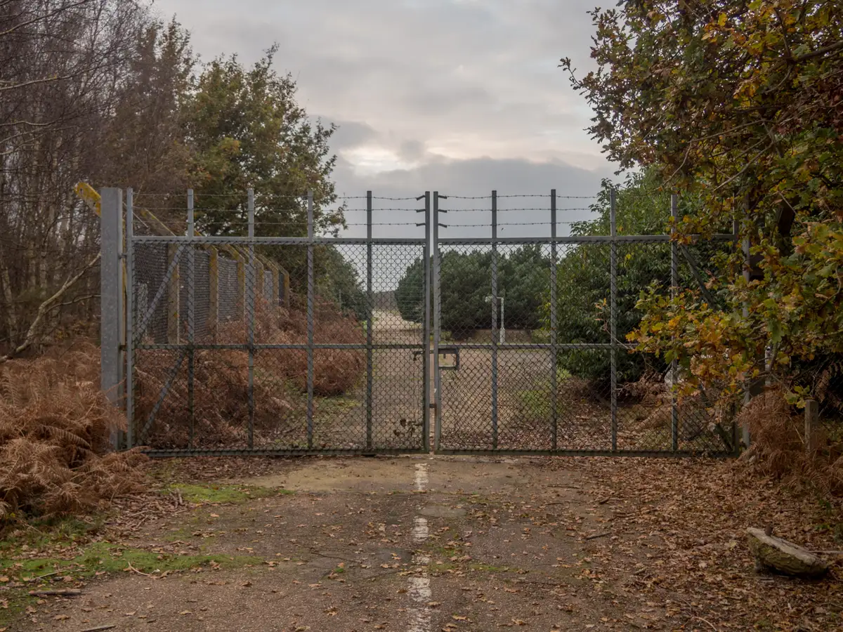 A closed metal mesh gate across a narrow concrete road, with a chain-link fence running off on each side between autumn trees and scrub.