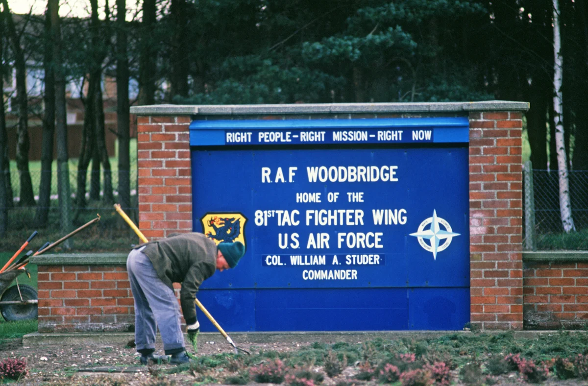 A blue sign on a brick frame reads RAF Woodbridge, home of the 81st Tac Fighter Wing, US Air Force, while a groundsman in overalls weeds the bed in front of it.