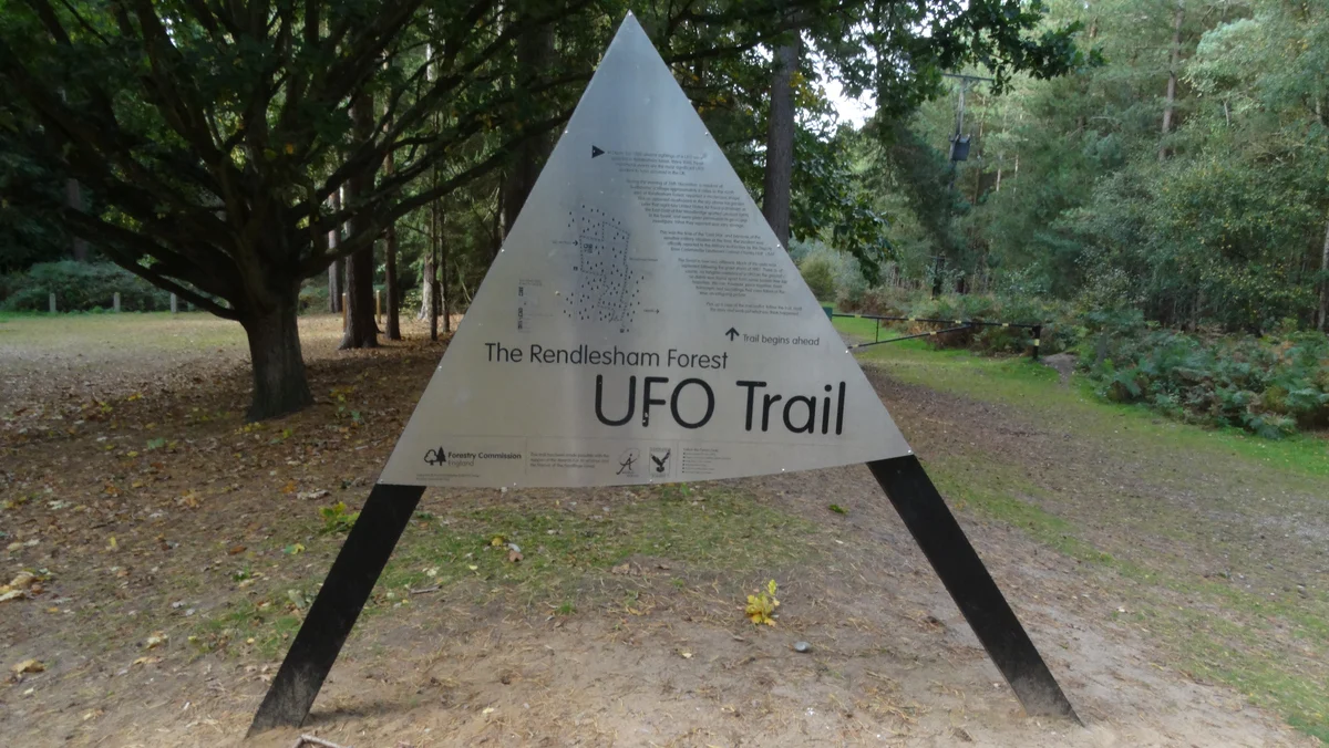 A triangular metal sign reading UFO Trail stands on a grassy clearing among pines and broadleaf trees in Rendlesham Forest.