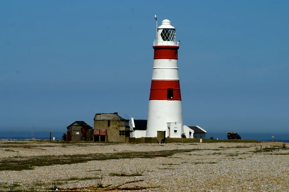 A red and white banded lighthouse tower with a white lantern room stands on a flat shingle spit beside low buildings, under a blue sky.