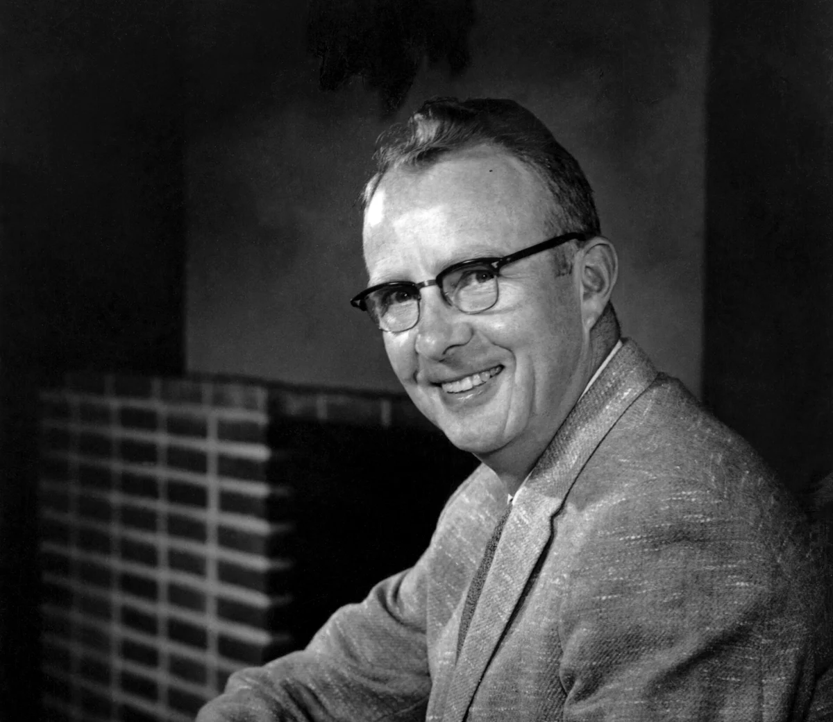 Black-and-white portrait of a smiling man in glasses, jacket and tie, seated in front of a dark background.