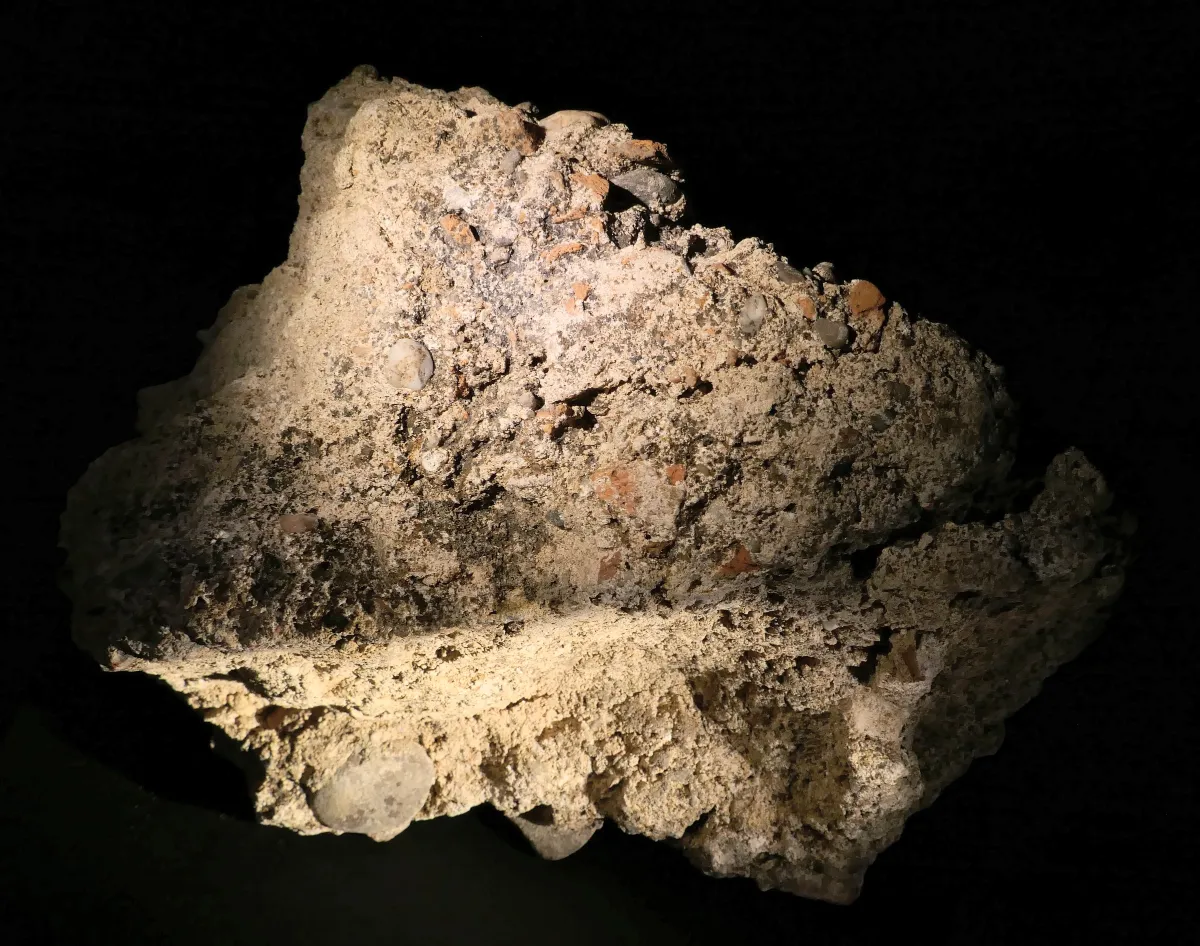 A broken lump of Roman concrete lit against black: rounded pebbles and pieces of brick set in pale, crumbly mortar.