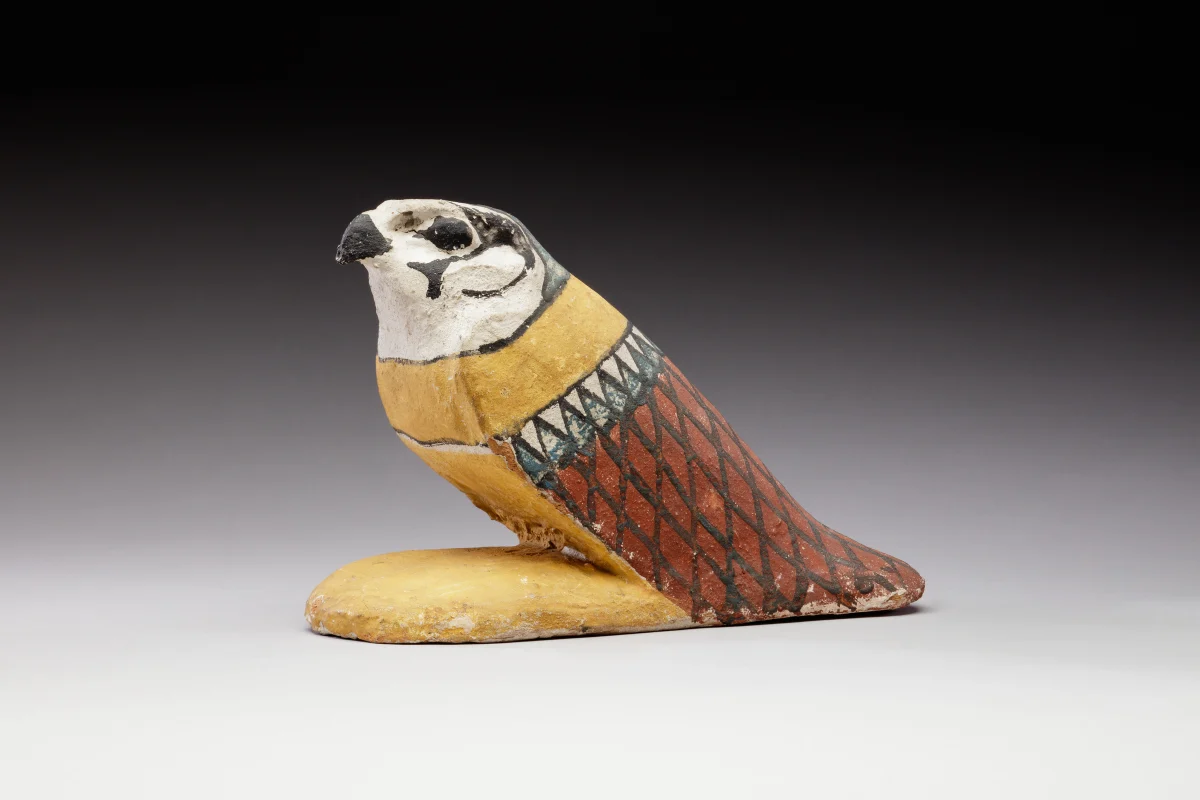 A painted wooden falcon sitting with folded wings, white face with dark markings, a yellow body and red feathers outlined in black.