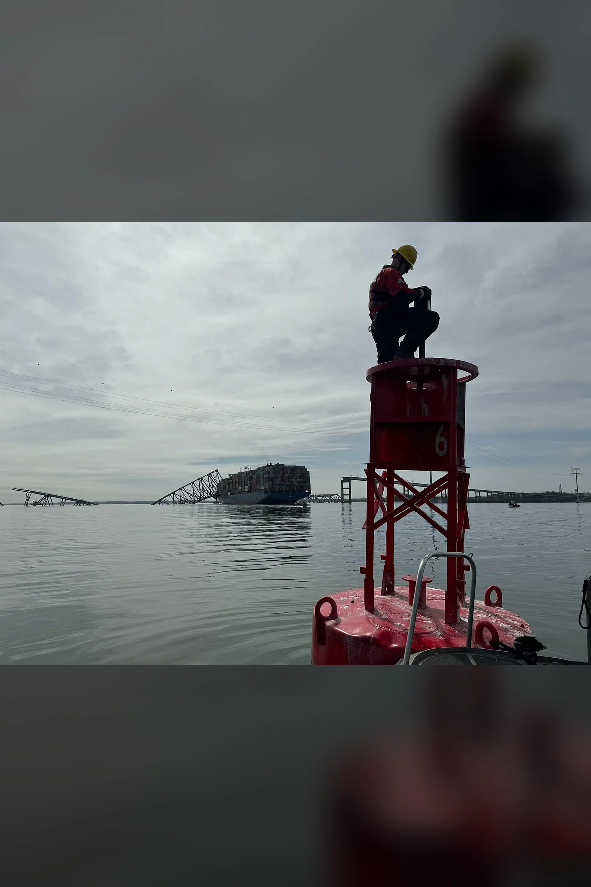 A worker on a red channel marker in grey water, the fallen bridge on the horizon.